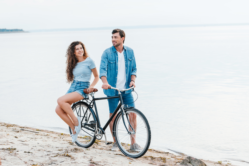 Man with bike and woman sitting at the back, next to the sea on the beach